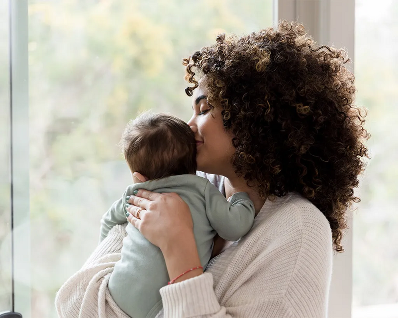 Eine Frau mit lockigem Haar hält ein Baby behutsam im Arm und gibt ihm einen liebevollen Kuss auf den Kopf.