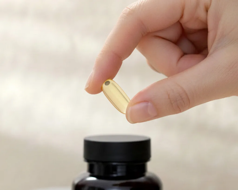 Eine Hand hält eine goldene, transparente Fischöl-Kapsel über eine Dose TISSO Pro Omega 3, die vor einem hellen, minimalistischen Hintergrund steht 