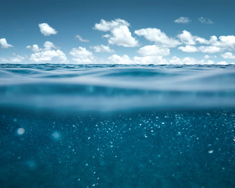 Meeresoberfläche in einer geteilten Ansicht: oben der weite blaue Horizont mit weißen Wolken, unten das tiefe, klare Wasser mit aufsteigenden Luftblasen