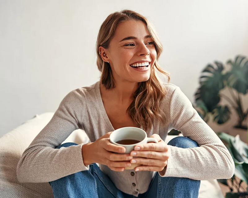 Lachende Frau mit Tee in der Hand sitzt auf dem Sofa